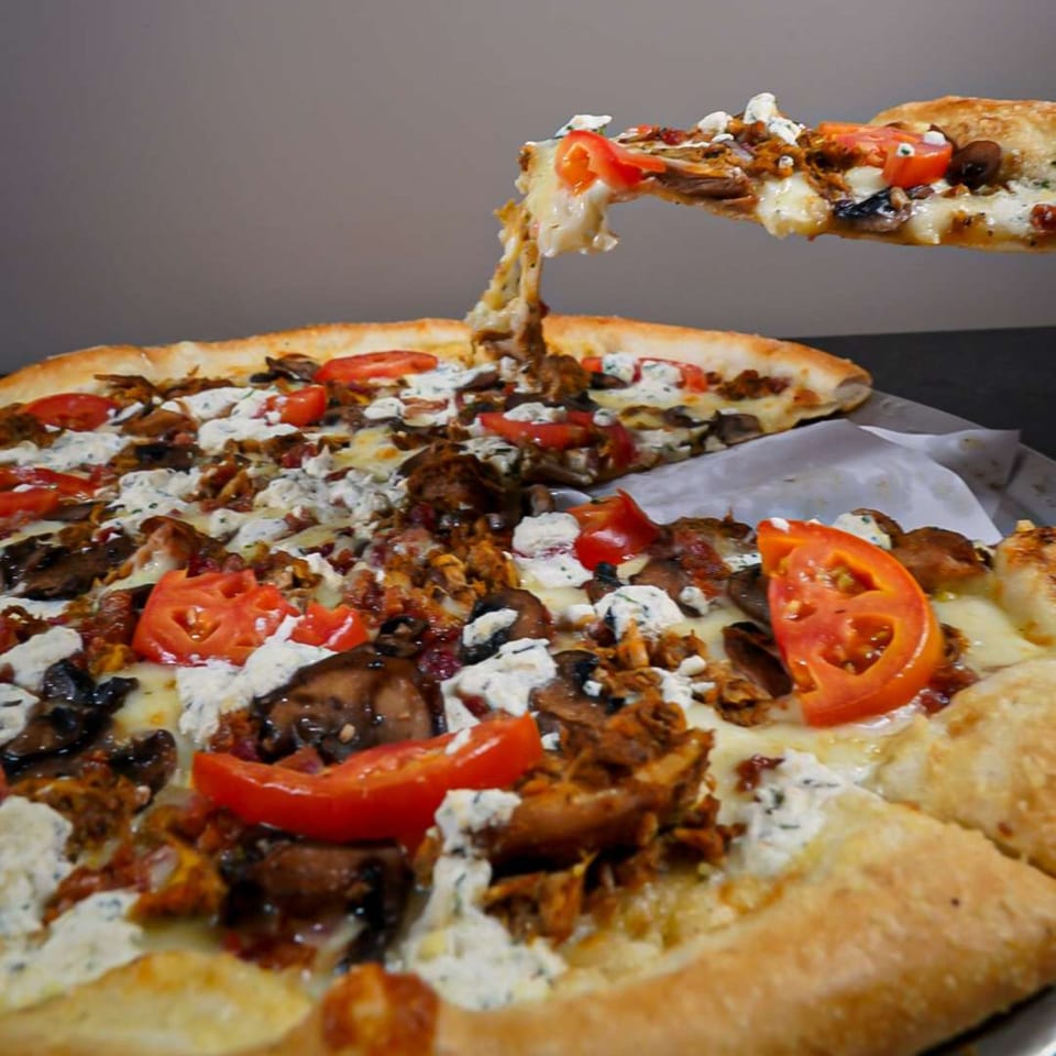 A slice of Ember + Ale pizza with melted cheese, tomatoes, and mushrooms being lifted from the pie.