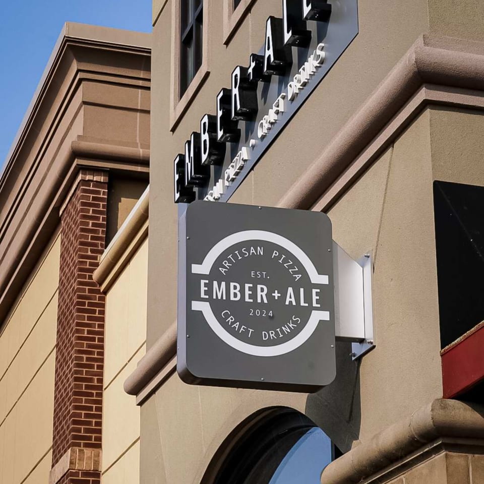 Ember + Ale restaurant exterior with a hanging sign and building-mounted letters against a tan facade.