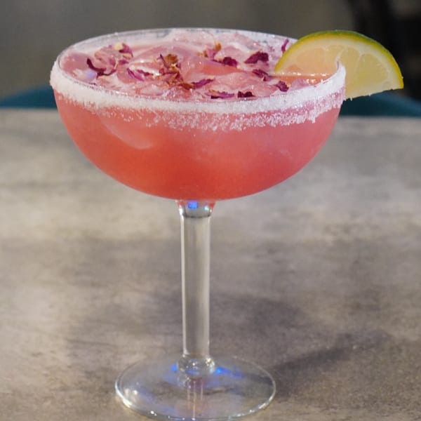 Pink cocktail in a margarita glass, garnished with dried rose petals and a lime wedge.