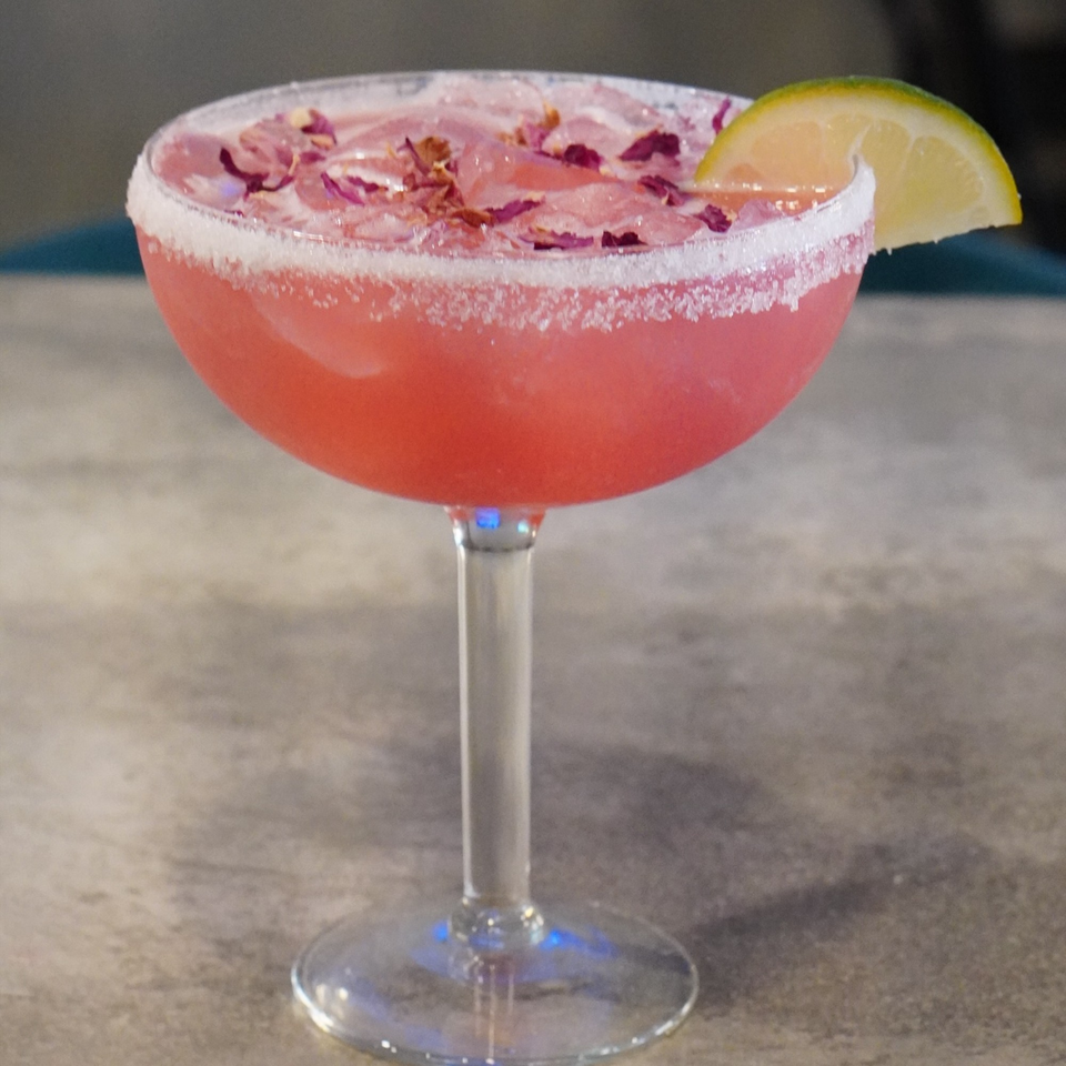 Pink cocktail in a margarita glass, garnished with dried rose petals and a lime wedge.