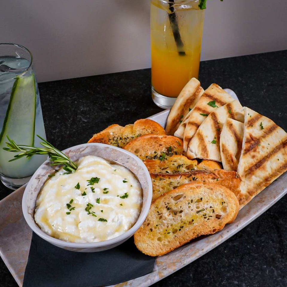Ember + Ale's baked ricotta dip with rosemary, served with toasted bread and grilled pita, alongside two drinks.