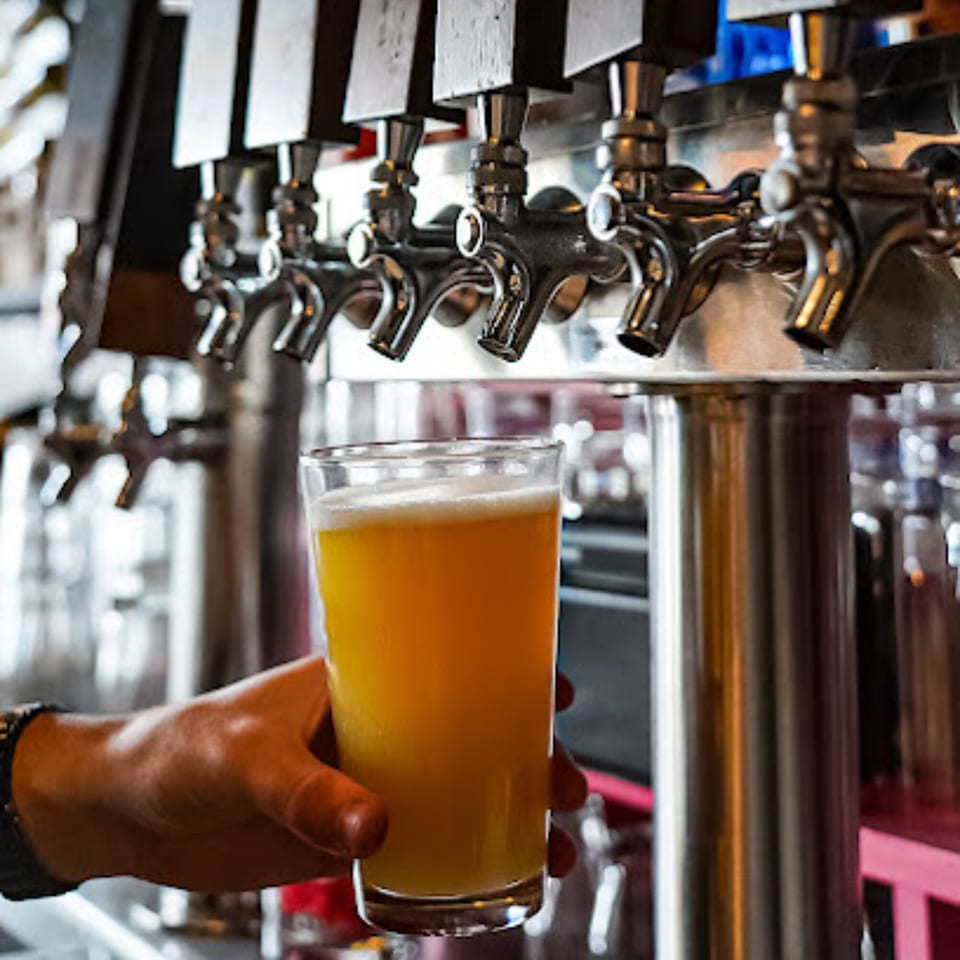 A hand holding a glass of Ember + Ale's golden beer, with beer taps in the background.