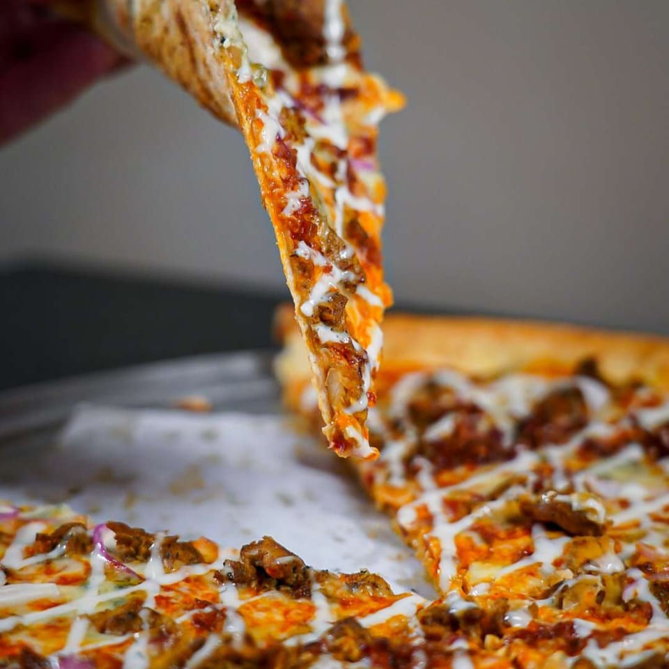 A hand lifts a slice of Ember + Ale pizza, topped with chicken, red onions, and white sauce.