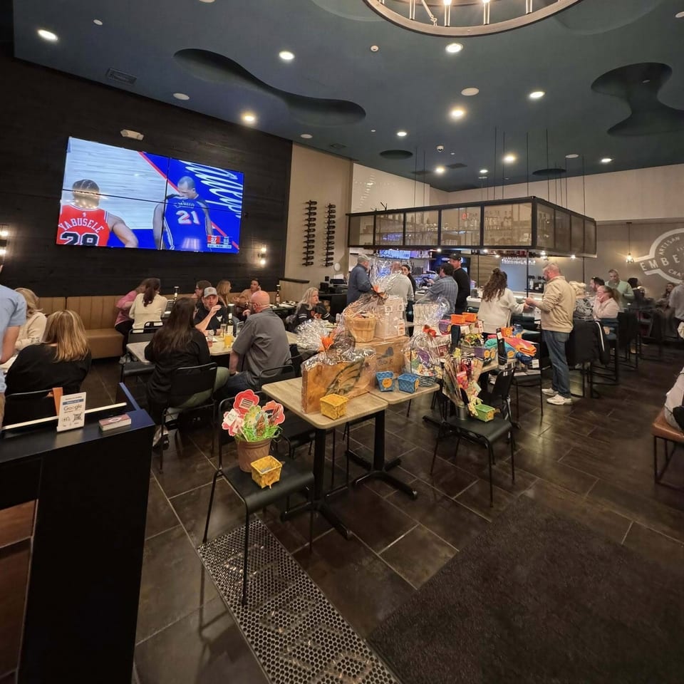A restaurant interior with patrons dining, a large TV screen showing basketball, and tables displaying gift baskets.