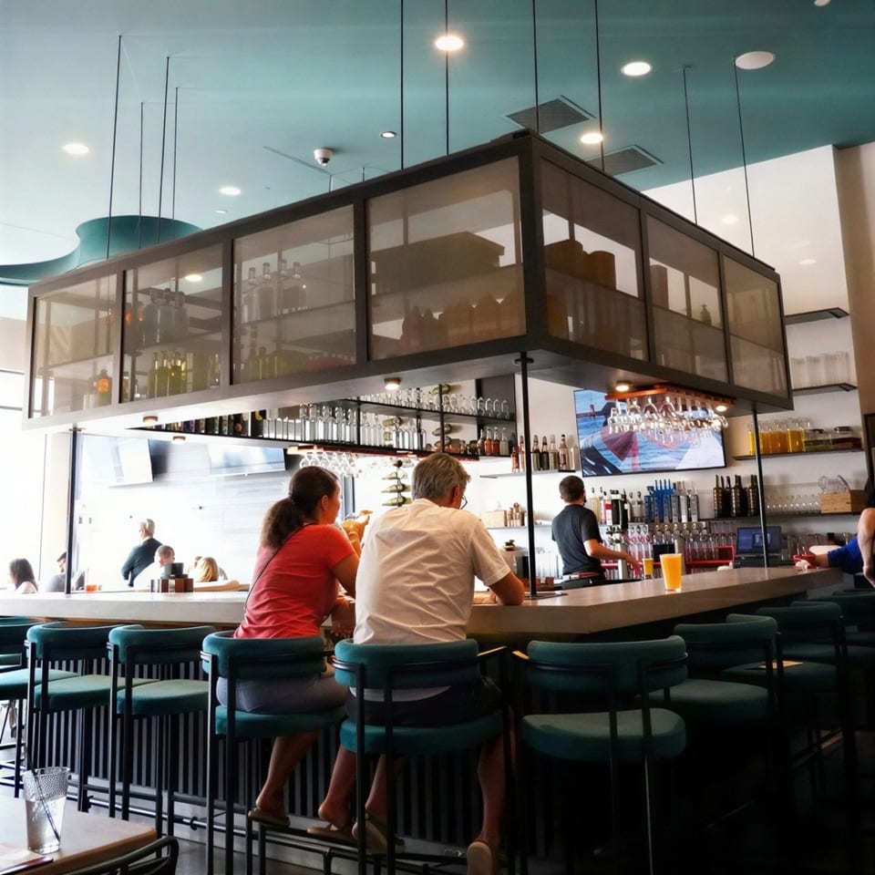 Patrons seated at a modern bar with teal stools, a bartender, and a TV screen.