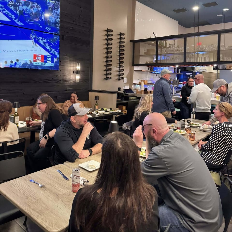 Diners seated at tables in a restaurant with a bar area and a large TV showing a basketball game.