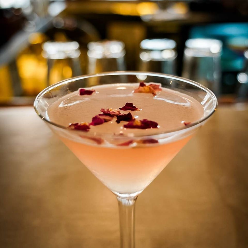 A pink martini with rose petals floating on top, set against a blurred bar background.
