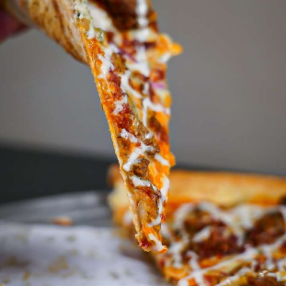 A hand holds up a slice of Ember + Ale pizza with melted cheese and toppings dripping.