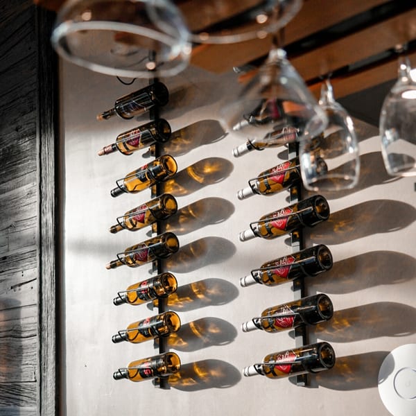 Wine bottles displayed on a wall rack with hanging wine glasses above, casting shadows.