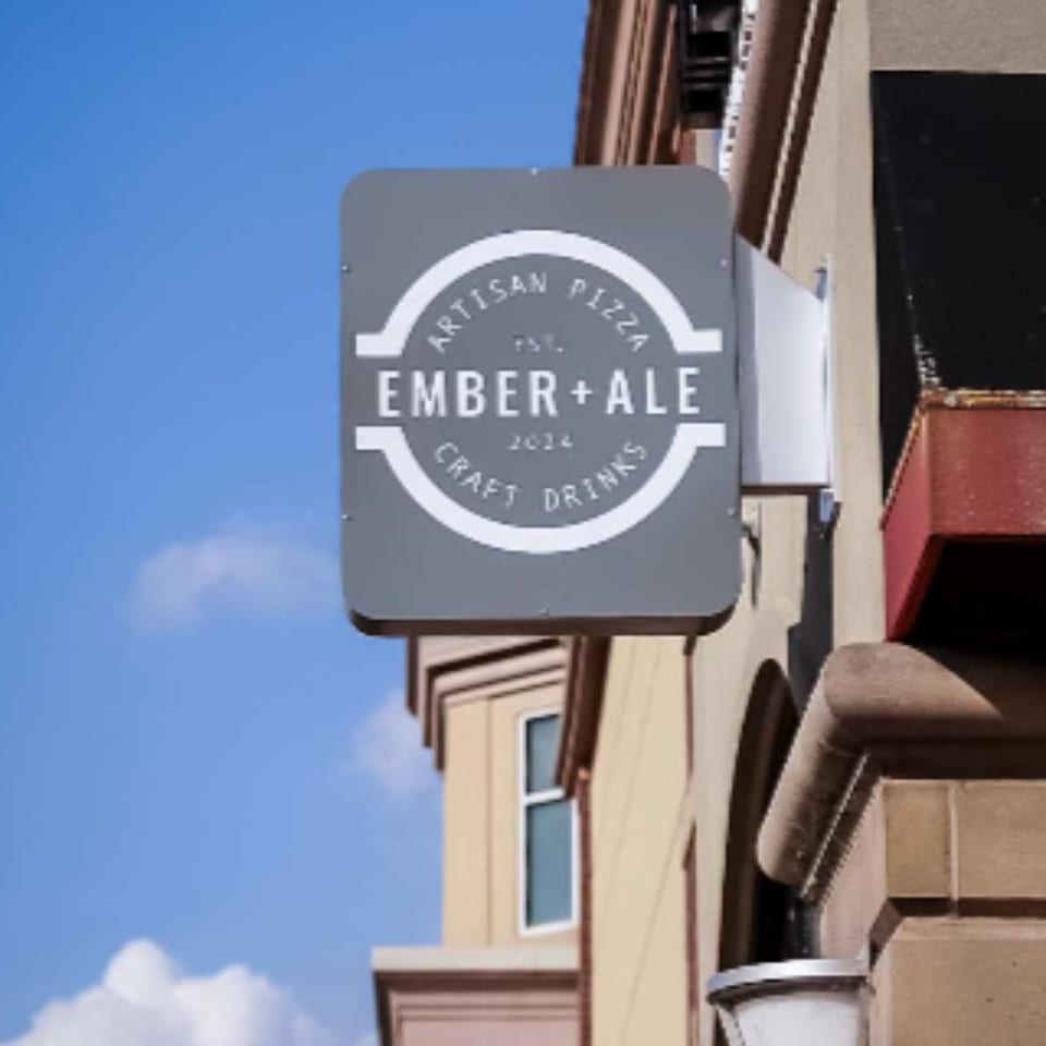 Ember + Ale artisan pizza and craft drinks sign, gray with white text, mounted on a building.