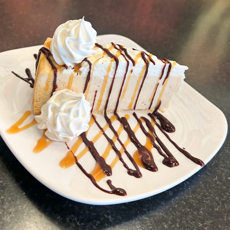 A slice of cheesecake with whipped cream, chocolate, and caramel drizzle on a white plate.