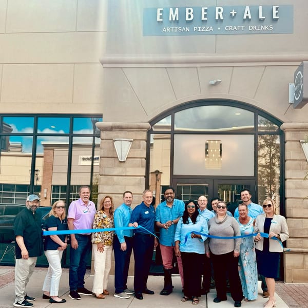 A group of people cutting a blue ribbon in front of Ember + Ale, an artisan pizza restaurant.