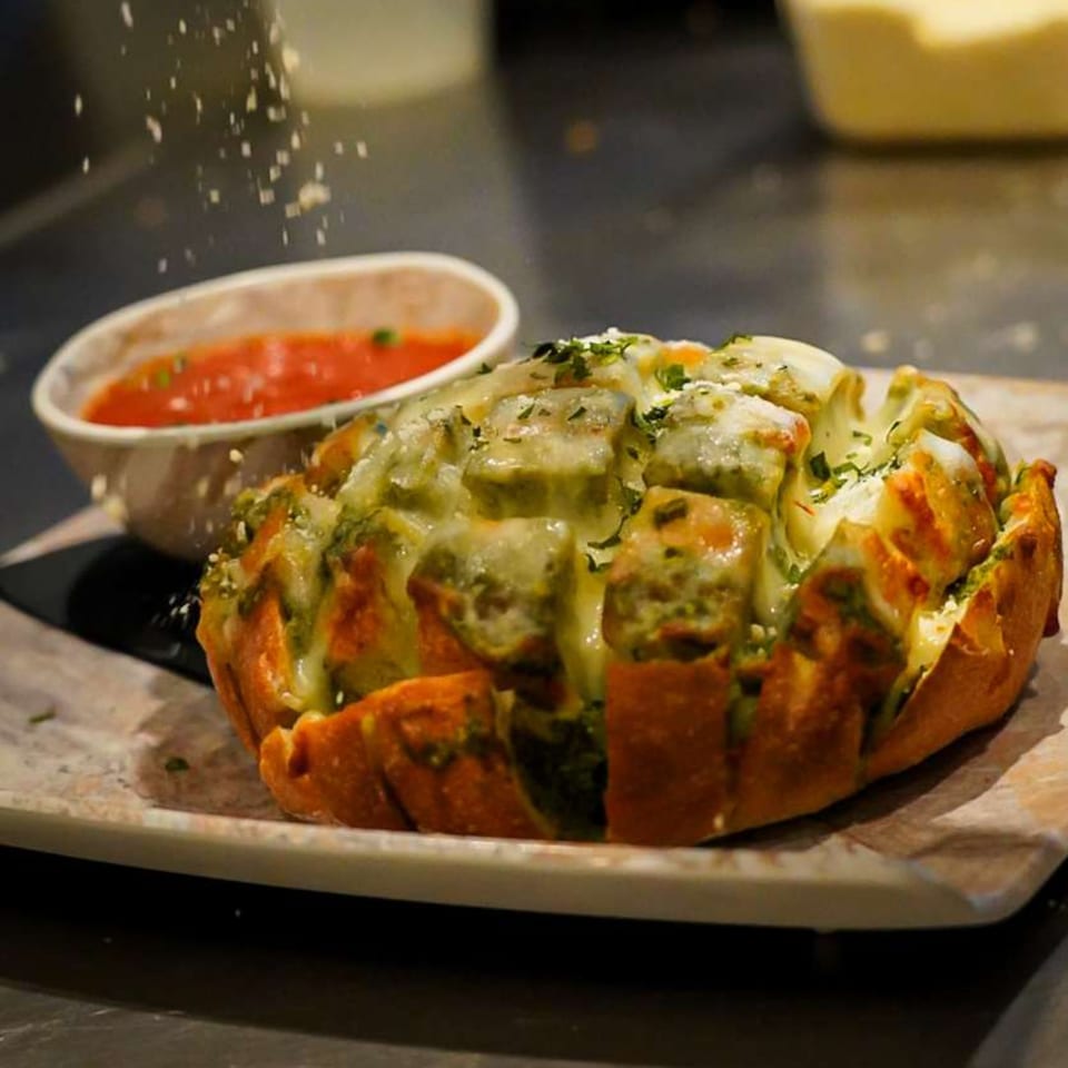 Pull-apart cheesy garlic bread with herbs, being sprinkled with cheese, next to a bowl of marinara.