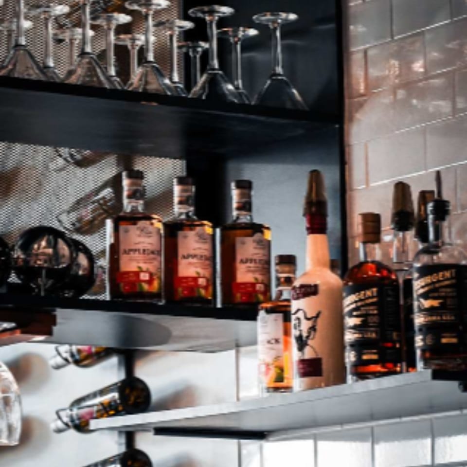Bar shelves at Ember + Ale displaying liquor bottles, wine, and hanging martini glasses.