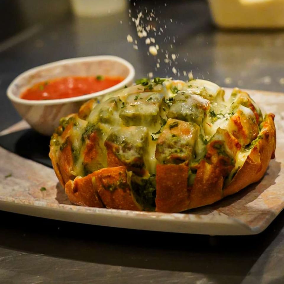 Cheesy pull-apart bread with green herbs, being sprinkled with cheese, next to a bowl of red sauce.