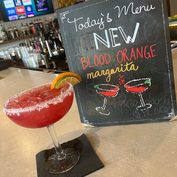 A red blood orange margarita with a lime and orange slice, next to a chalkboard menu.